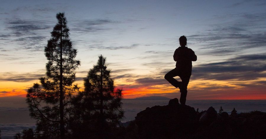 Silhouette of a practitioner in tree pose on a rock at sunset above a sea of clouds