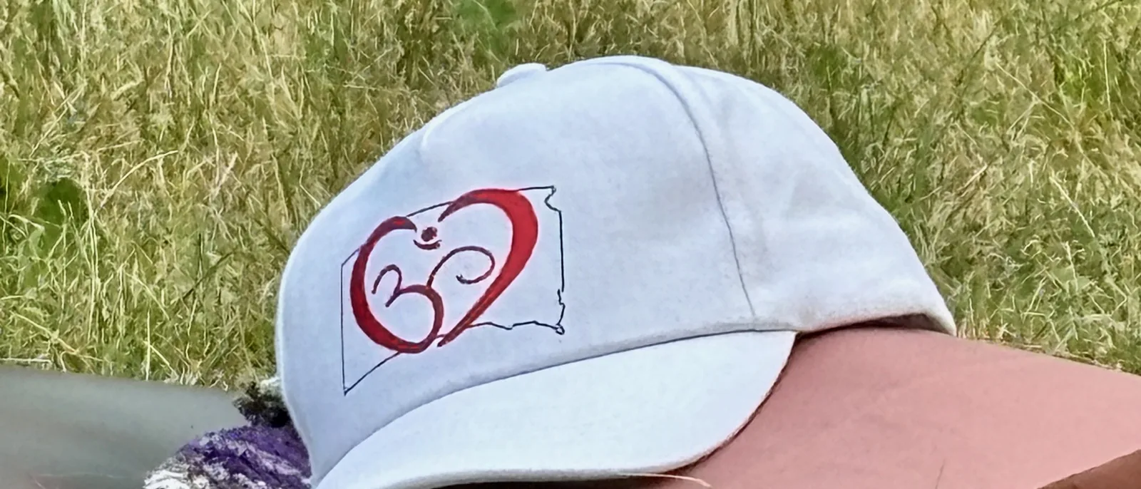 Festival cap with the heart-and-om South Dakota mark resting on the meadow grass