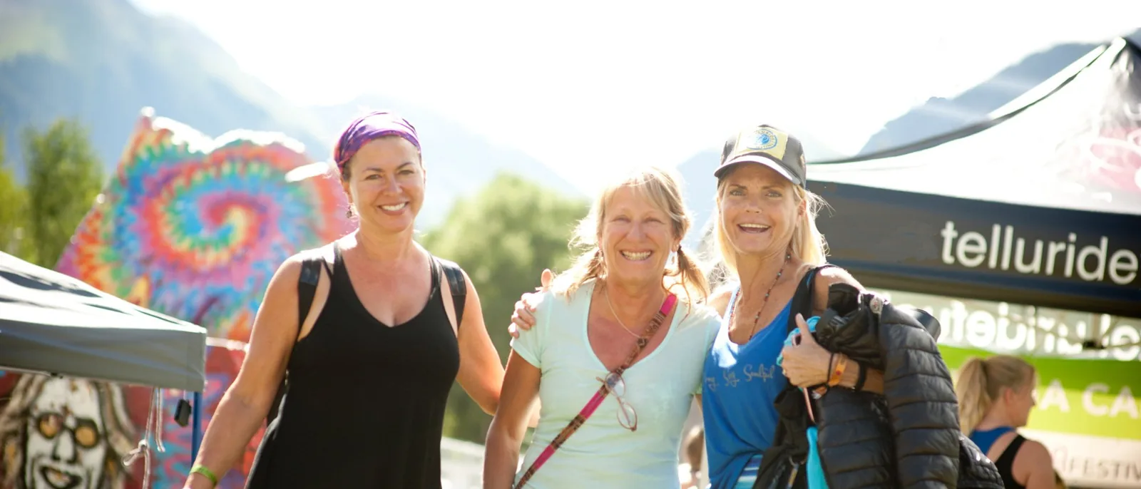 Three friends arm in arm at the Telluride Yoga Festival village, tents and peaks behind