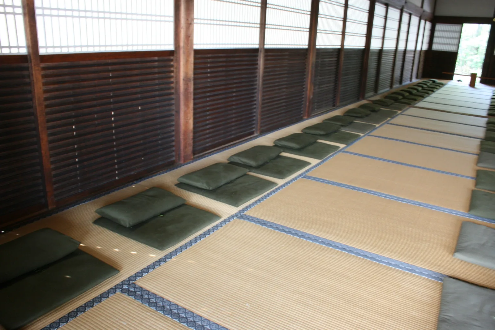 Rows of flat green cushions laid out along a tatami floor in a long, quiet wooden hall with slatted screens.