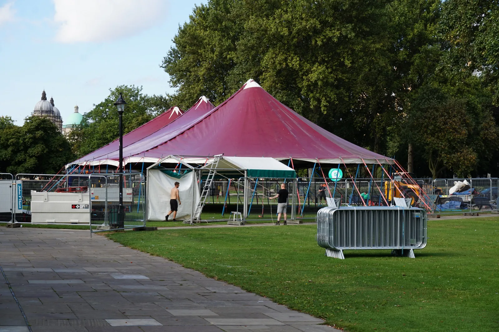 A red and white peaked marquee tent pitched on a lawn in a public park, with trees and a domed building behind it.