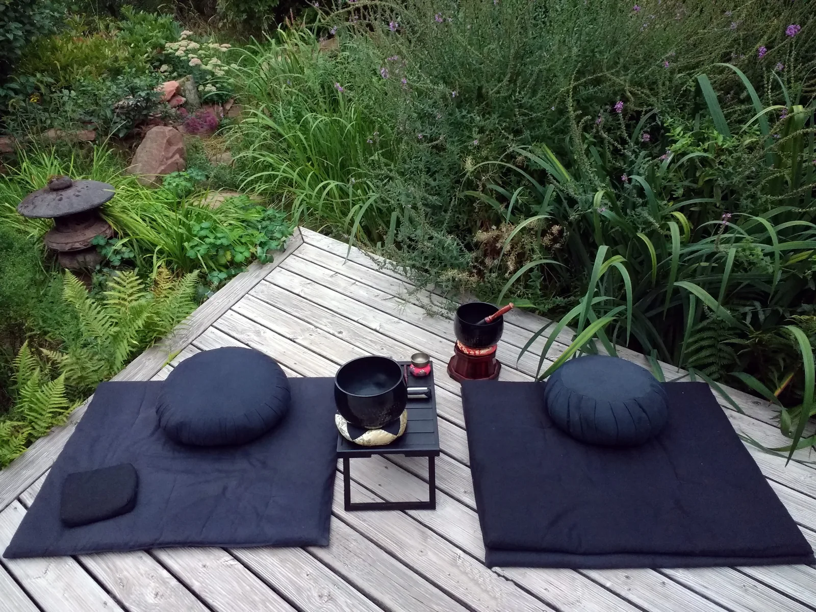 Two dark round meditation cushions and flat mats laid out on a wooden deck beside tall grasses, with a low table and small bowls between them.