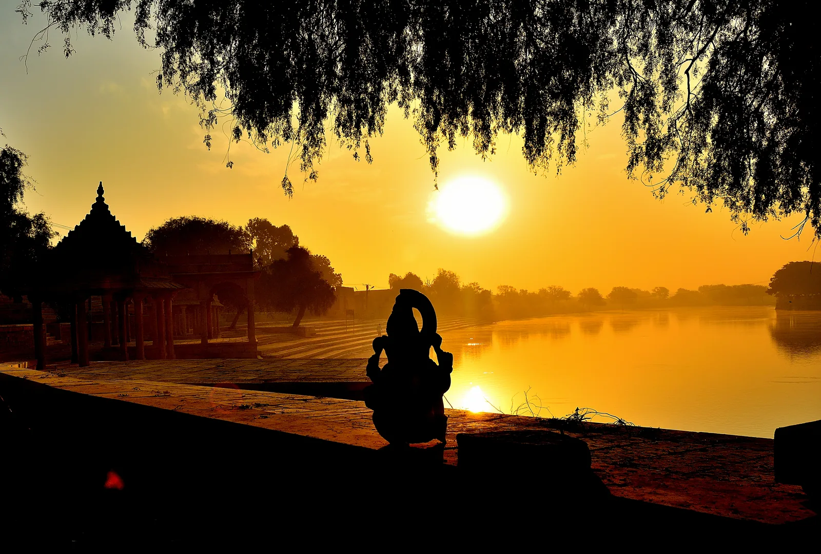 A silhouetted figure sitting cross-legged on the stone bank of a lake at sunrise, with a domed pavilion to one side.