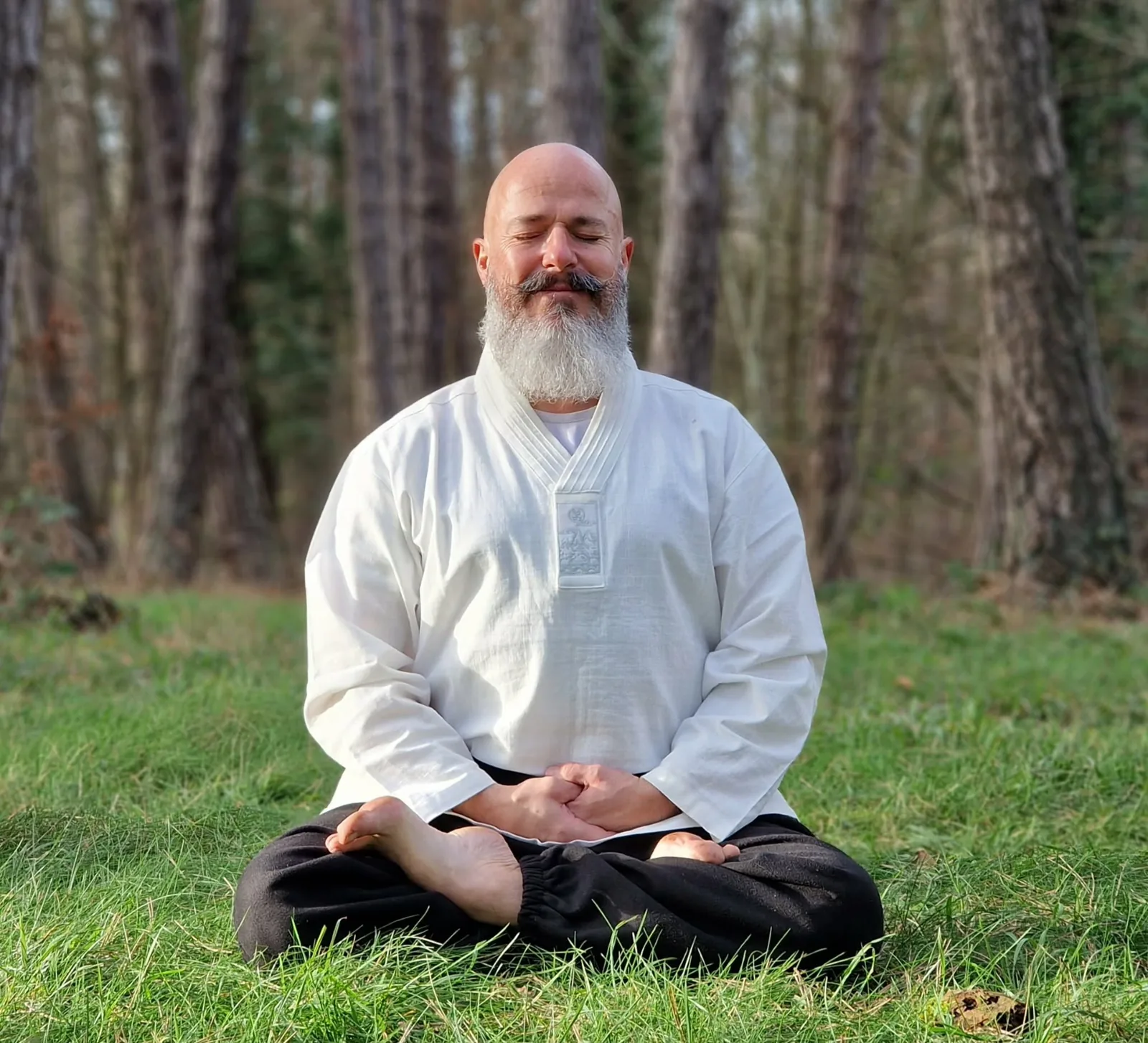 A bearded man sitting cross-legged on grass in light woodland, hands resting on his knees and eyes closed.