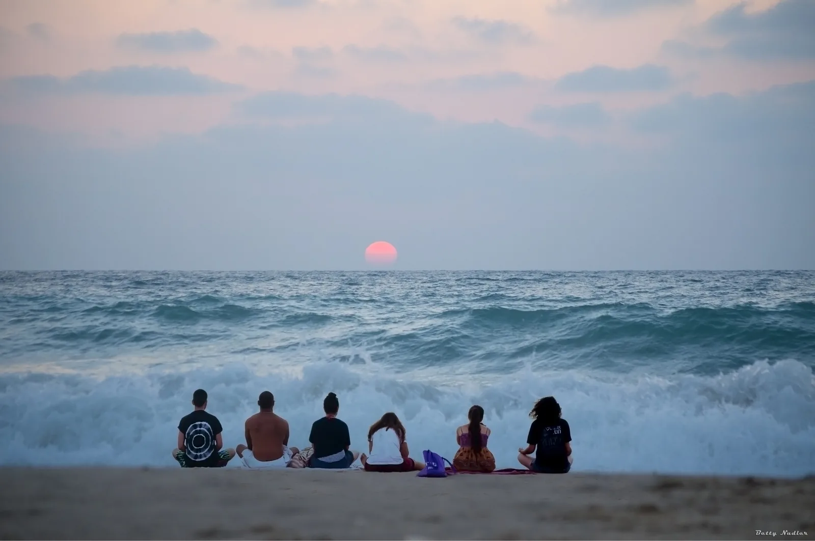 Eight people sitting cross-legged in a row on wet sand, facing a breaking wave and a low sun over the sea.