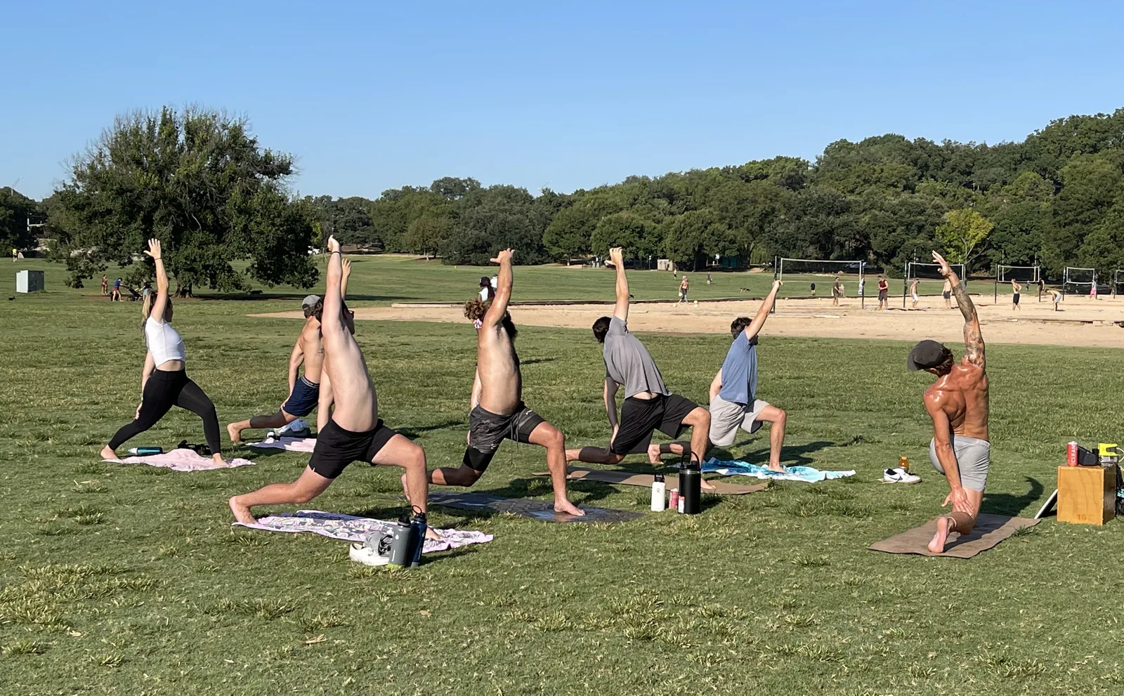 A row of people on mats holding a standing side stretch on a park lawn, with open grass and trees behind them.