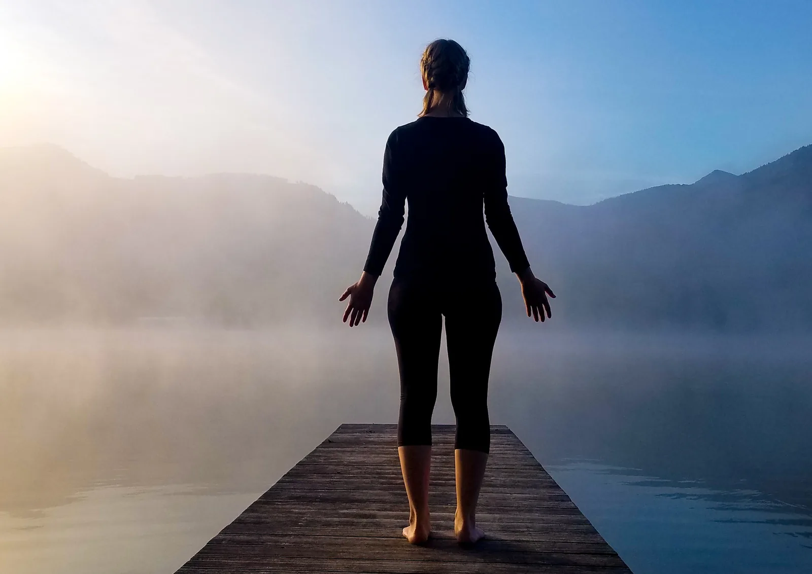 A person standing at the end of a wooden jetty at dawn, arms low and open, facing mist over a calm lake and hills.