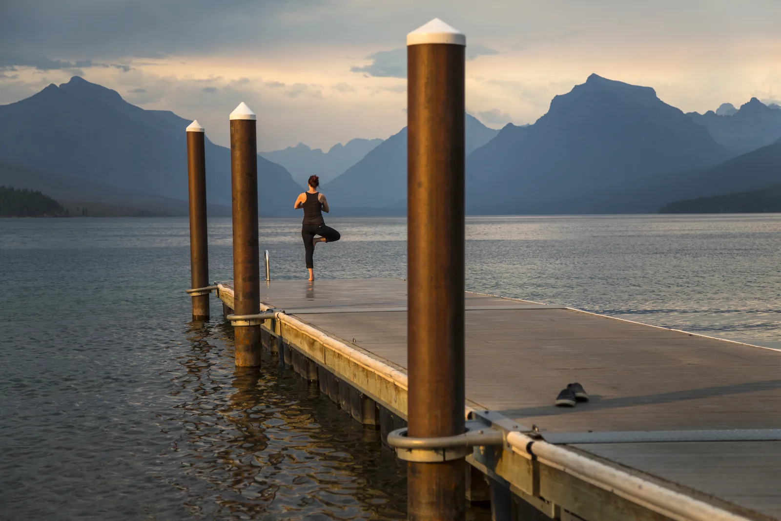 A distant figure standing in tree pose at the end of a wooden lake jetty, with mountains and still water behind.