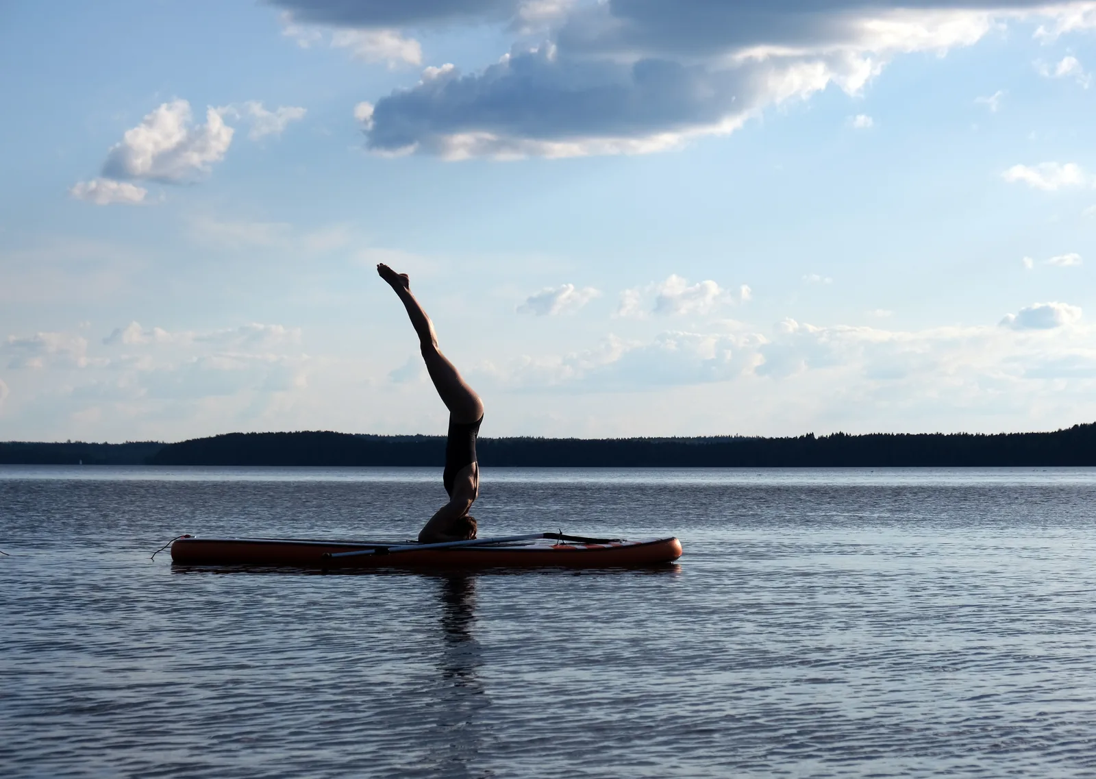 A person holding a headstand on a paddleboard in the middle of a calm lake under a blue sky with scattered cloud.