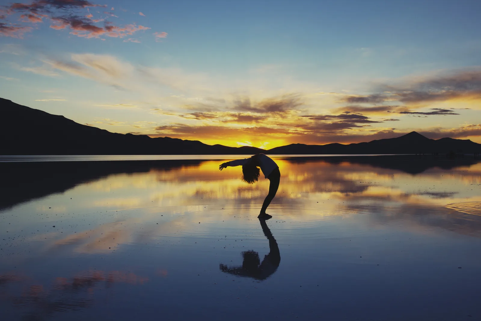 A person arching backwards on a mirror-like flooded salt flat, their reflection doubled beneath them under a broad sky at sunset.