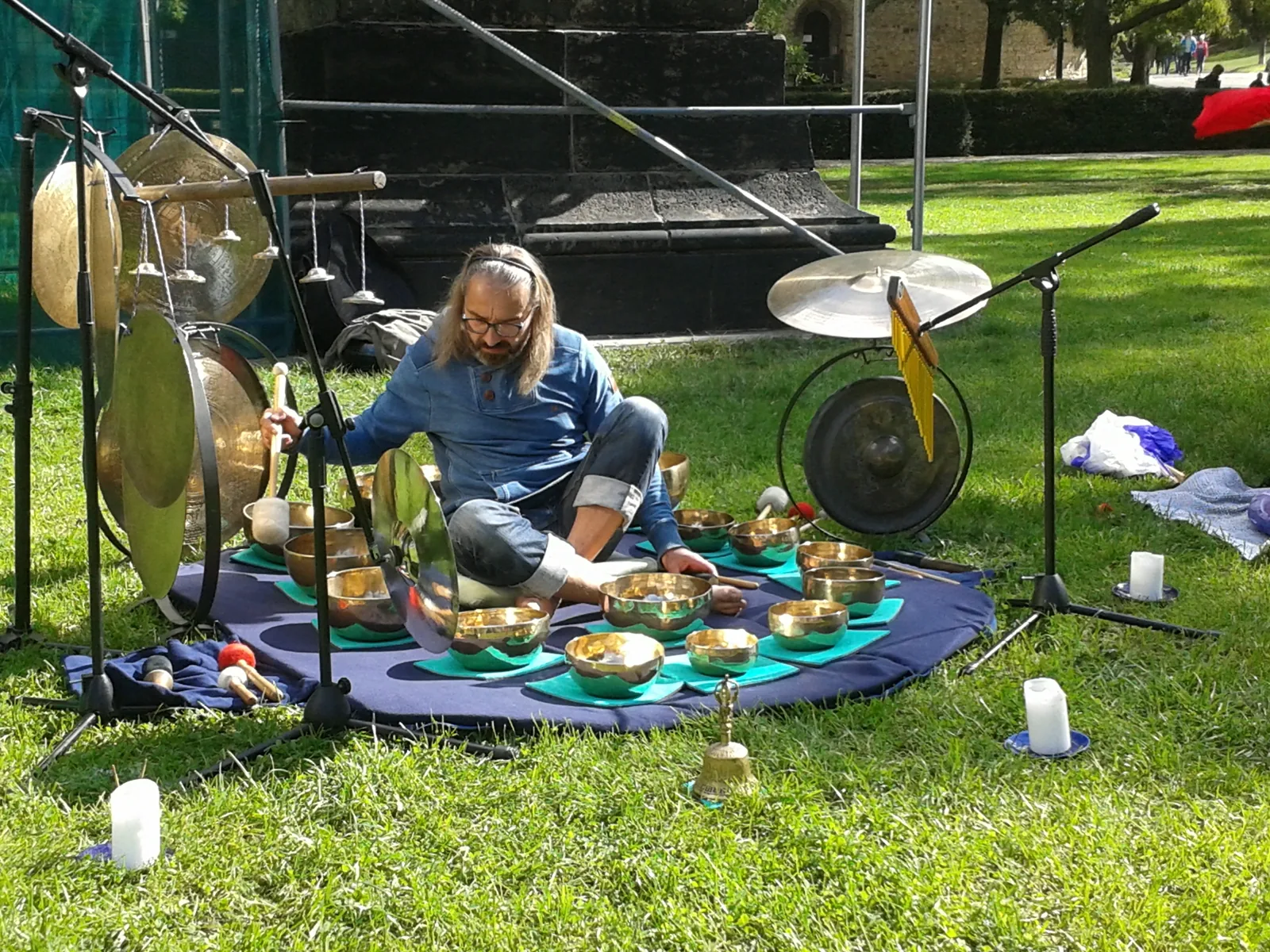 A person seated on a rug on grass surrounded by hanging gongs, metal singing bowls and candles, playing mid-session.