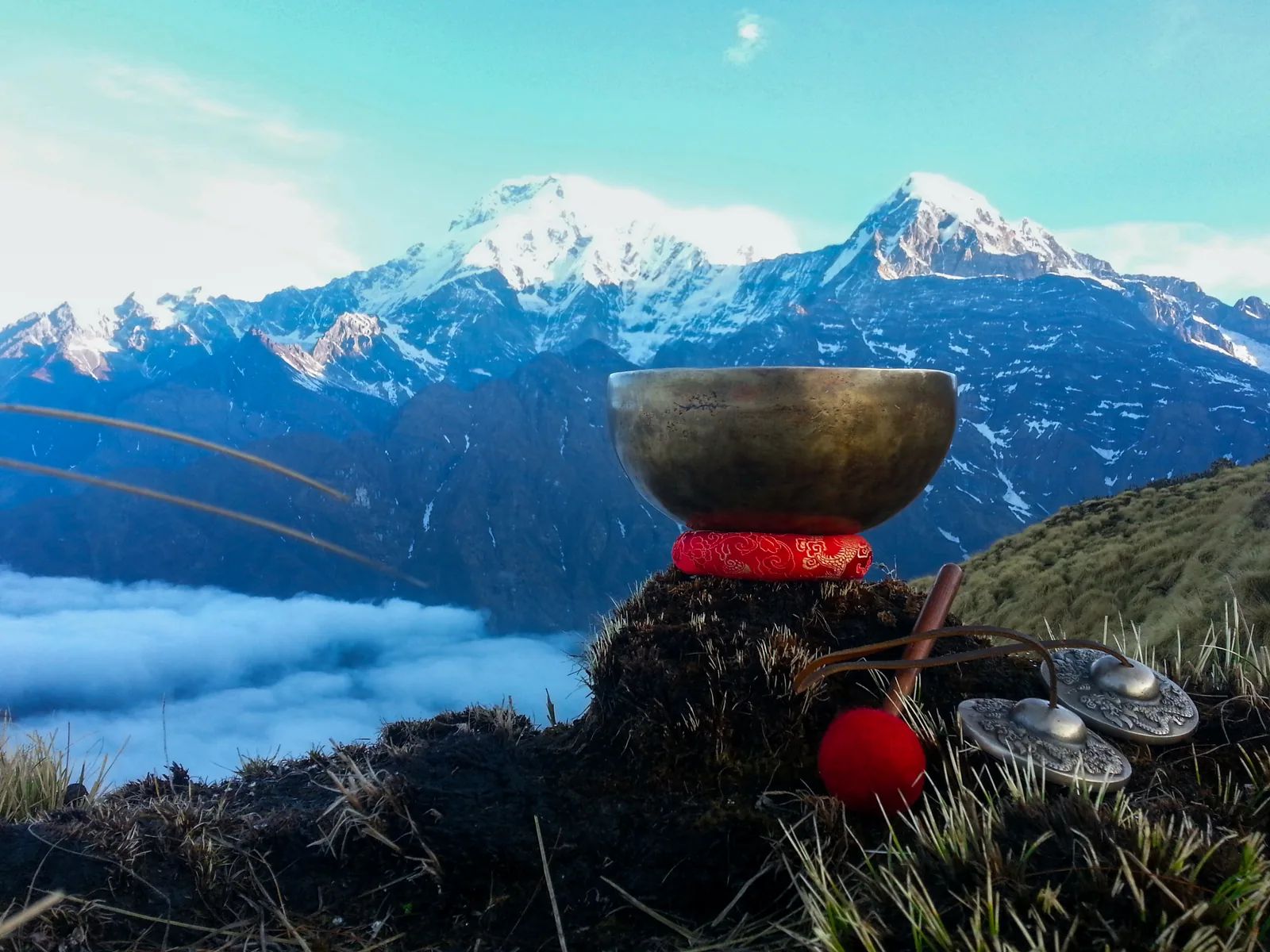 A metal singing bowl and striker resting on a red cushion on a rock, with snow-covered mountain peaks and cloud in the distance.